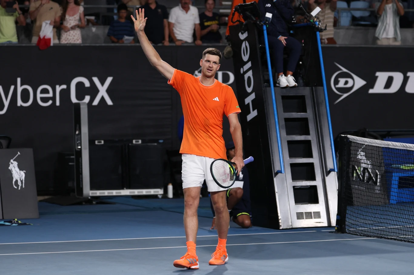 Hubert Hurkacz tenisista w pomarańczowej koszulce i białych spodenkach stoi na korcie tenisowym, trzyma rakietę w jednej ręce i unosi drugą dłoń w geście powitania lub podziękowania, w tle widać sędziego na podwyższeniu oraz kibiców na trybunach