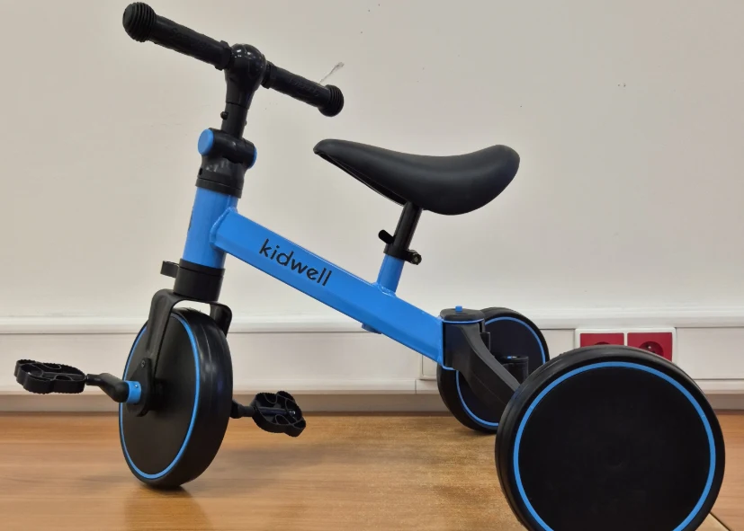 niebiesko-czarny dziecięcy rowerek trójkołowy z napisem „kidwell” na ramie, ustawiony na drewnianej podłodze na tle białej ściany