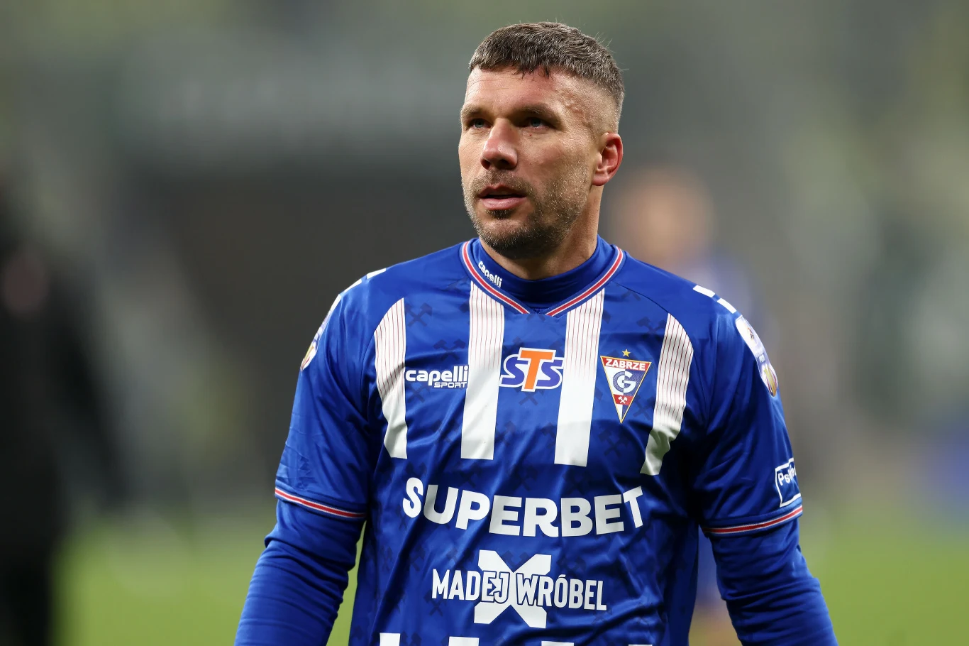 piłkarz w niebieskiej koszulce z białymi pasami, z herbem klubu i logo sponsorów, stojący na boisku podczas meczu; wyraz twarzy skupiony, w tle rozmyte trybuny i murawa