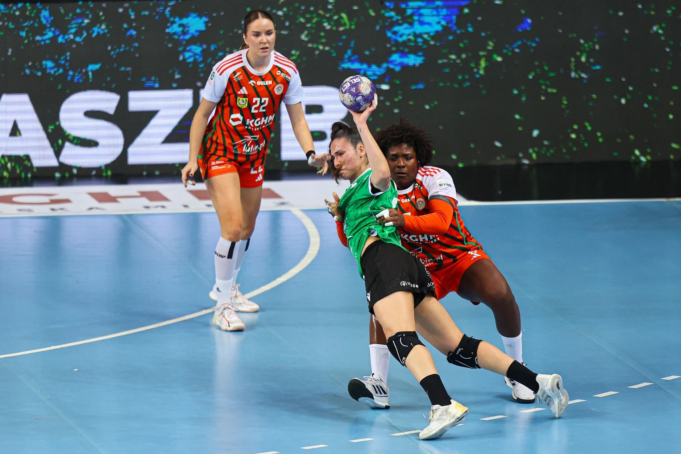 dynamiczna akcja podczas meczu piłki ręcznej kobiet, zawodniczka w zielonej koszulce próbuje oddać rzut do bramki, będąc ściśle broniona przez rywalkę w pomarańczowym stroju; w tle kolejna zawodniczka z tej samej drużyny obserwuje sytuację