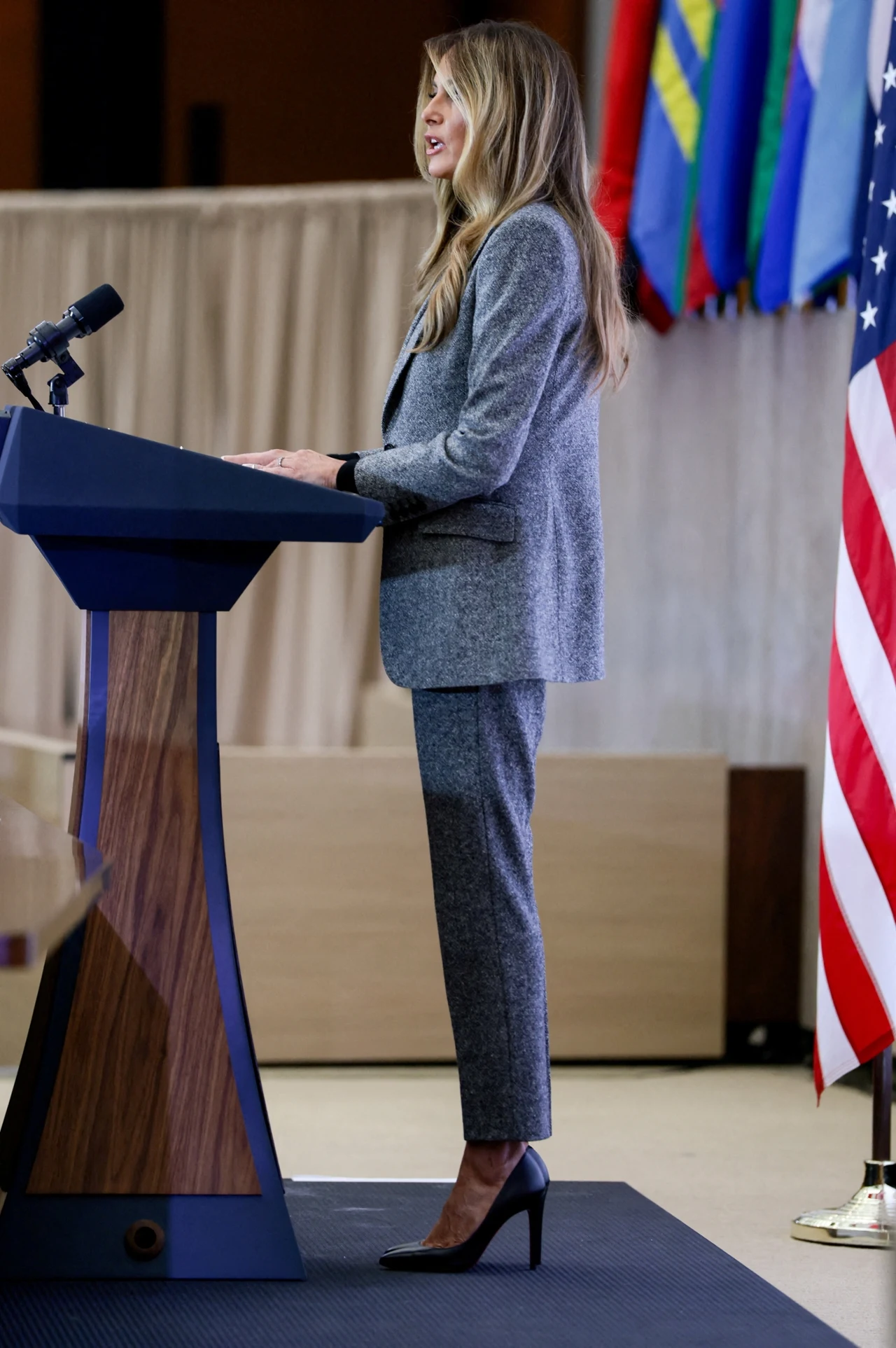Kobieta w szarym garniturze i czarnych szpilkach stoi przy mównicy, wygłaszając przemówienie; w tle widoczne są kolorowe flagi oraz flaga Stanów Zjednoczonych.