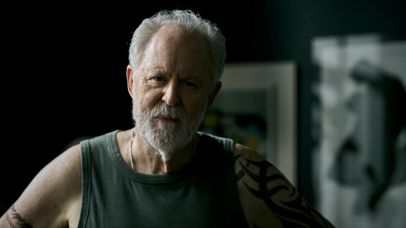 starszy mężczyzna z siwą brodą i tatuażem na ramieniu, ubrany w ciemny podkoszulek, stoi w półcieniu na tle rozmytych obrazów w galerii lub pracowni