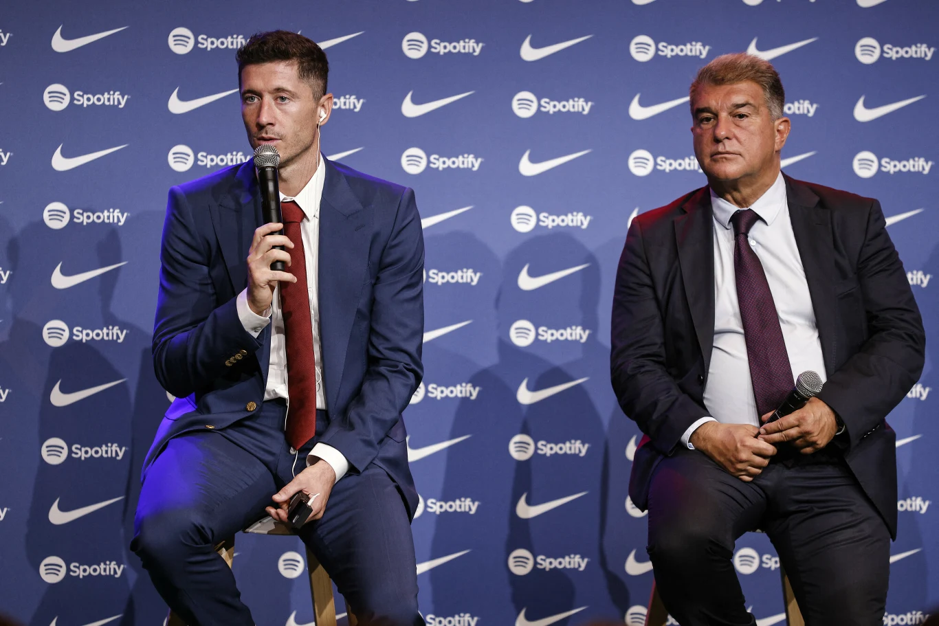 Dwaj mężczyźni w eleganckich garniturach siedzą na wysokich krzesłach na tle ścianki z logotypami Spotify i Nike; jeden z nich trzyma mikrofon i przemawia, drugi słucha z poważnym wyrazem twarzy, trzymając mikrofon na kolanach.