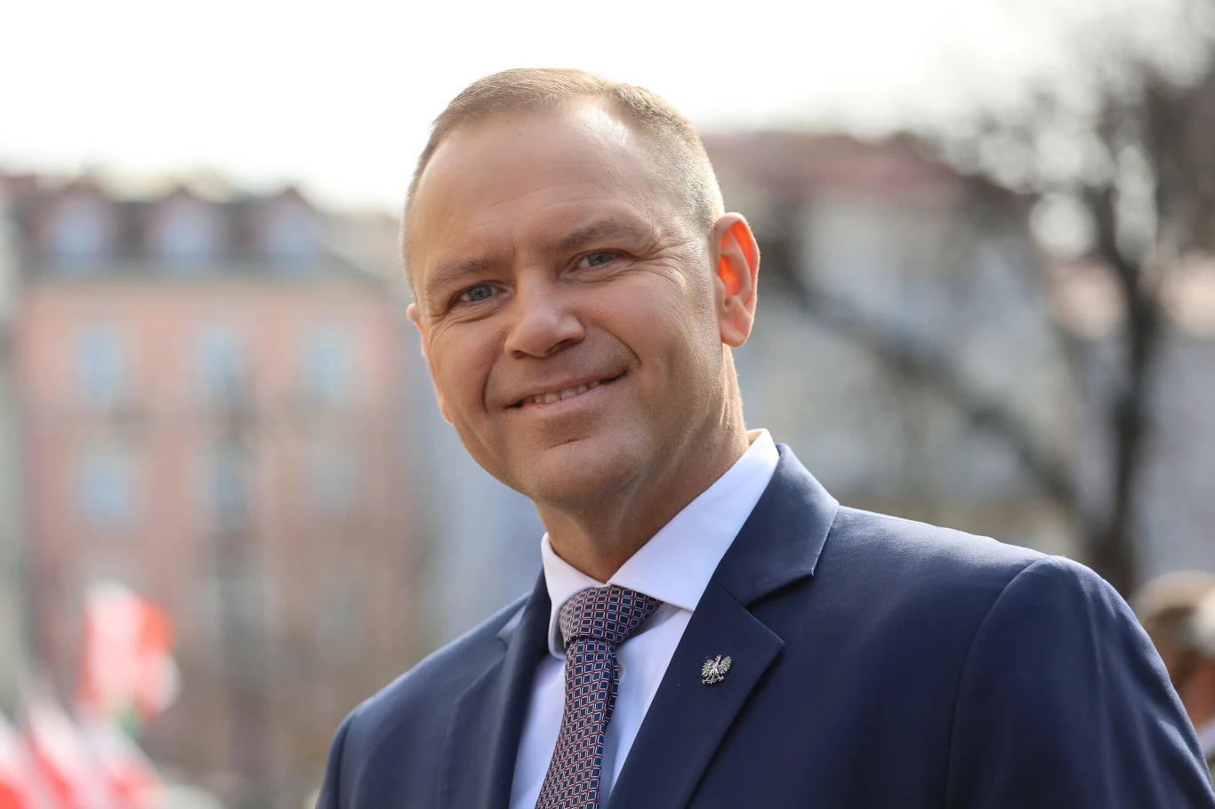 Mężczyzna w garniturze i krawacie uśmiecha się, stojąc na tle rozmytego miejskiego krajobrazu z kolorowymi budynkami i delikatnym światłem dziennym.