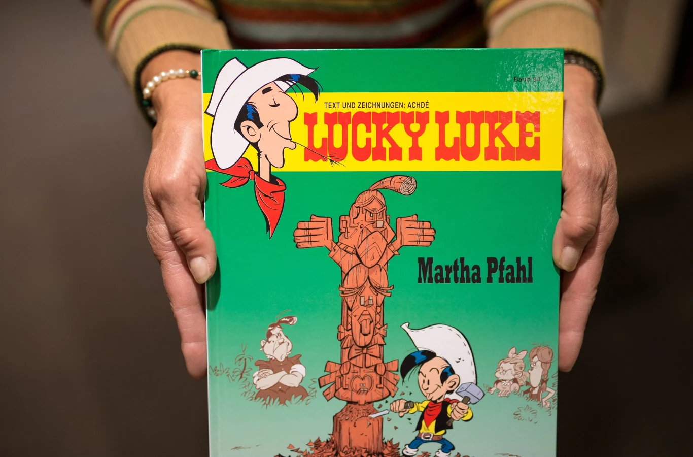 Komiks Lucky Luke trzymany oburącz przez osobę w swetrze, na okładce widoczny kolorowy rysunek głównego bohatera z białym kapeluszem oraz totemem; z lewej strony postacie z komiksu, na górze wyraźny żółty tytuł na zielonym tle.