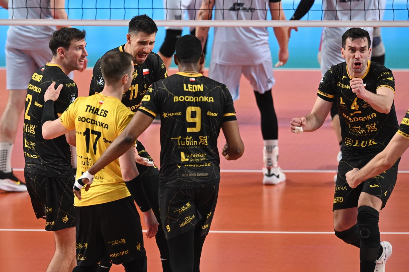 Grupa siatkarzy w czarno-żółtych strojach celebruje zdobyty punkt na boisku podczas meczu siatkówki; w tle widoczni są przeciwnicy w jasnych koszulkach za siatką.