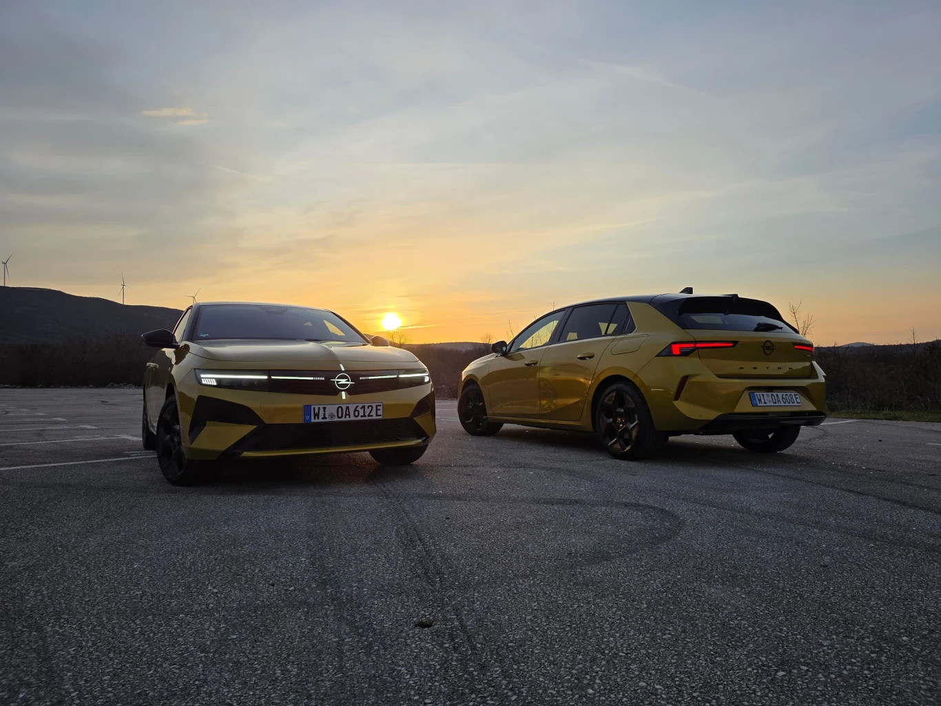 Dwa żółte samochody marki Opel ustawione na asfalcie, jeden skierowany przodem, drugi tyłem, na tle zachodzącego słońca oraz gór widocznych na horyzoncie.