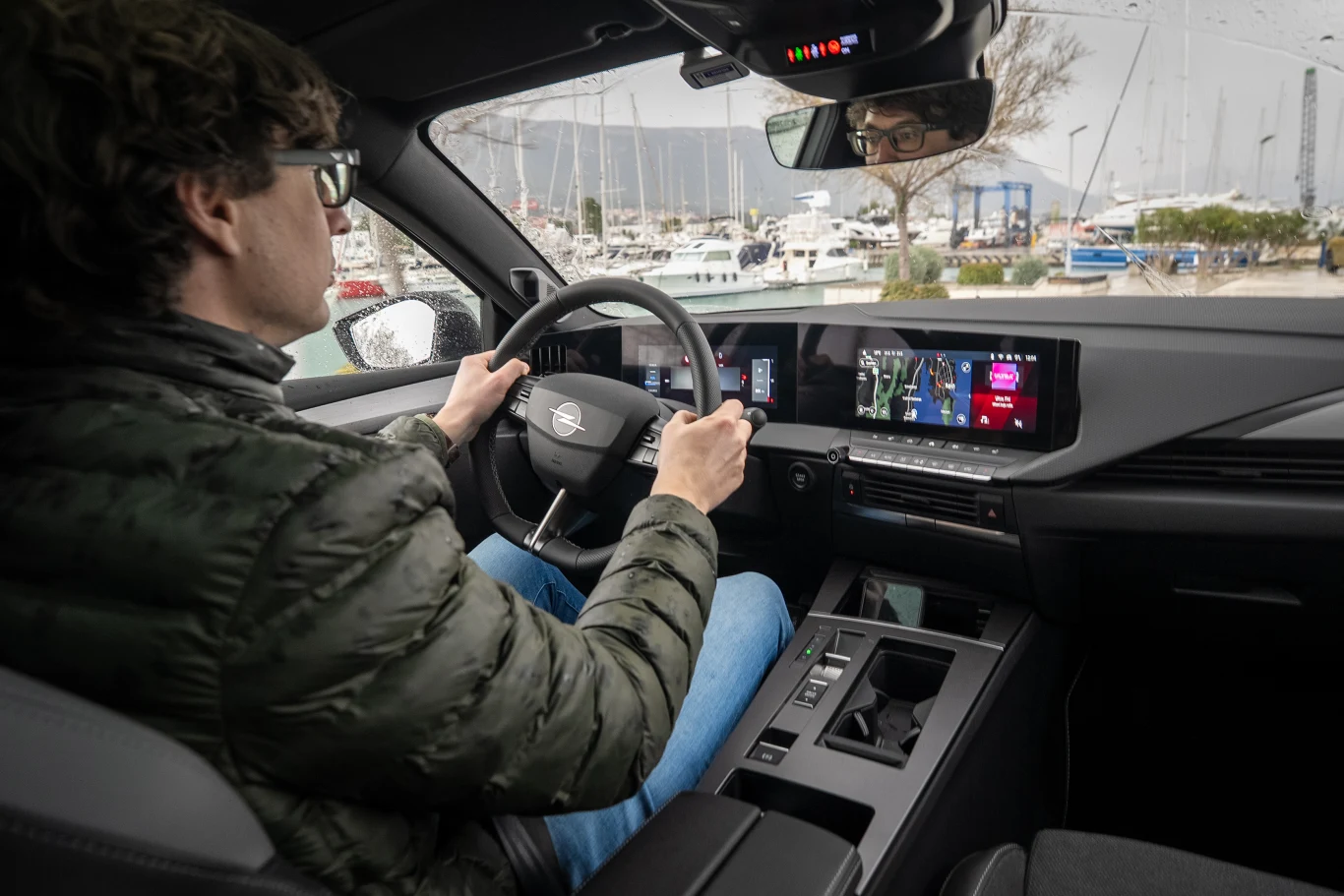 mężczyzna w okularach prowadzi nowoczesny samochód z cyfrowym kokpitem i ekranem nawigacji, na zewnątrz widoczna marina z jachtami