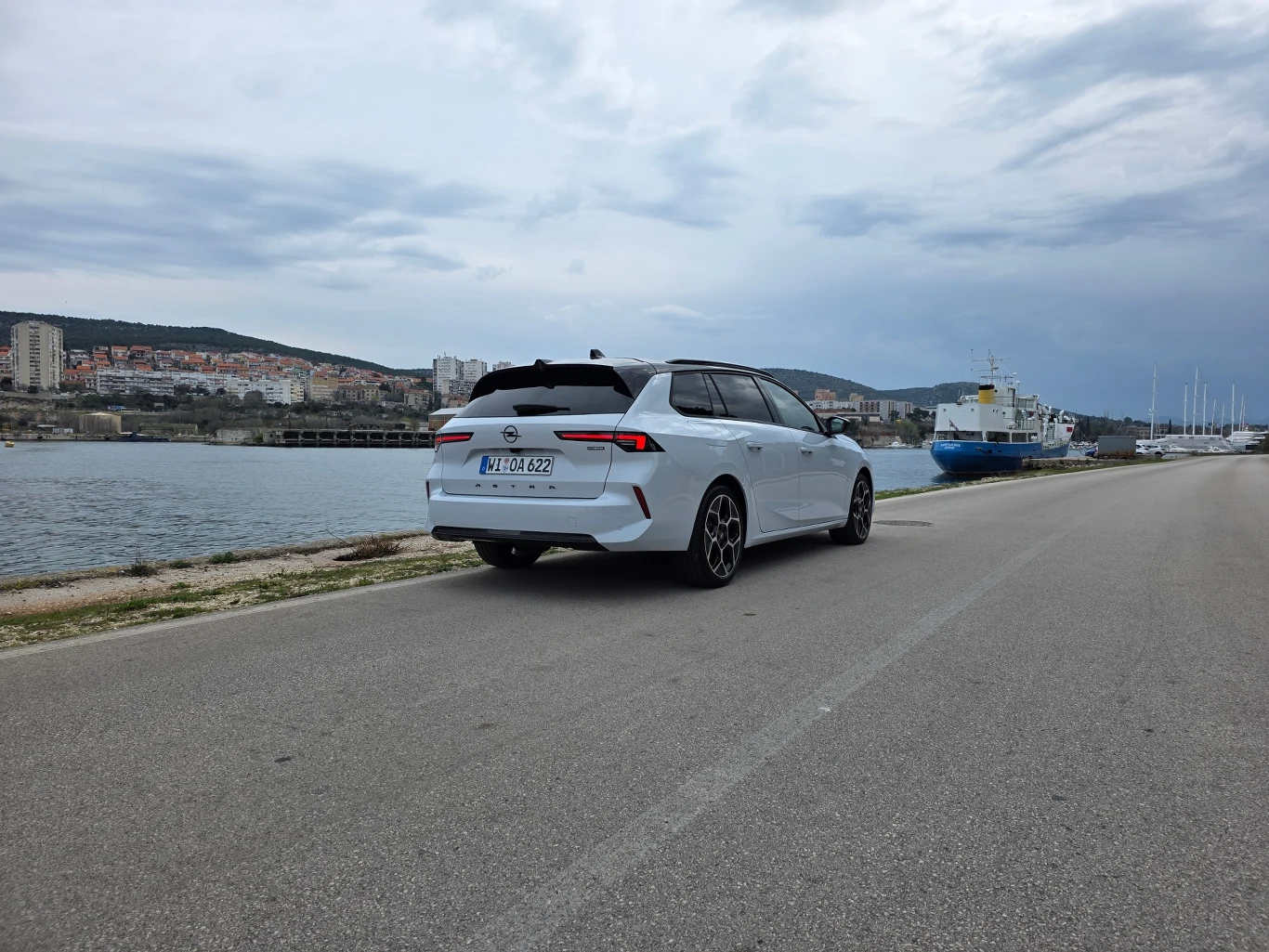 Białe kombi zaparkowane nad wodą przy szerokiej, pustej drodze, w tle widoczne miasto na wzgórzach oraz kilka zacumowanych statków na nabrzeżu.