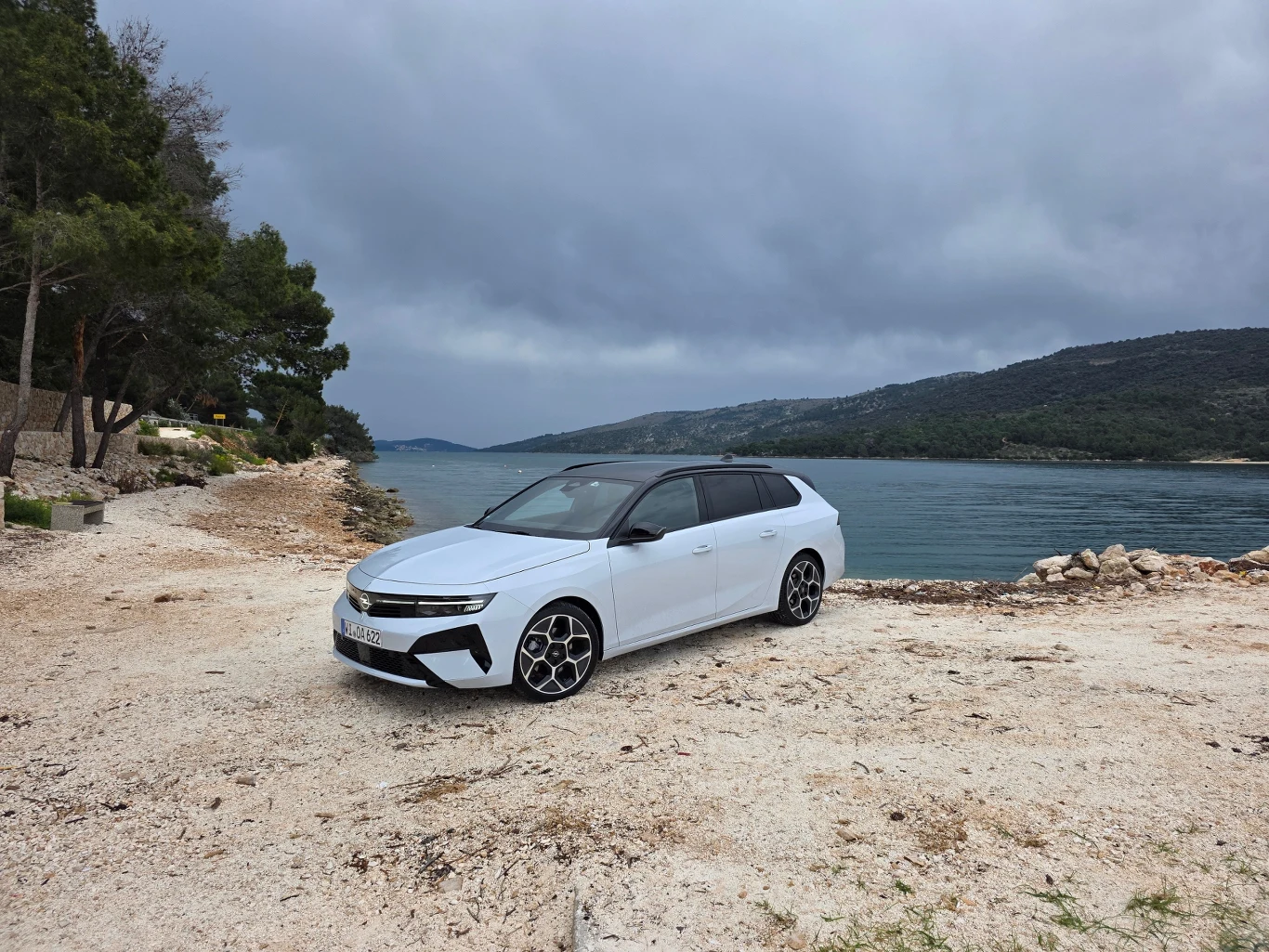 biały samochód typu kombi stoi na żwirowym brzegu w pobliżu spokojnej wody, w tle widoczne są zielone wzgórza oraz pochmurne niebo