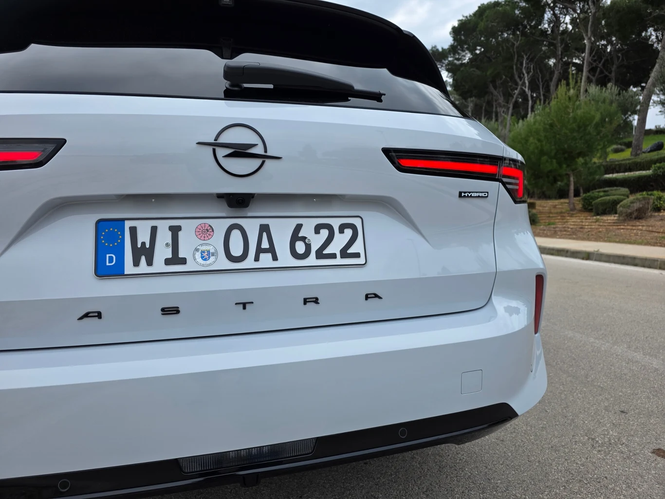 biały Opel Astra z niemiecką tablicą rejestracyjną, nowoczesnym tylnym oświetleniem LED i oznaczeniem modelu na klapie bagażnika, zaparkowany przy ulicy z drzewami w tle