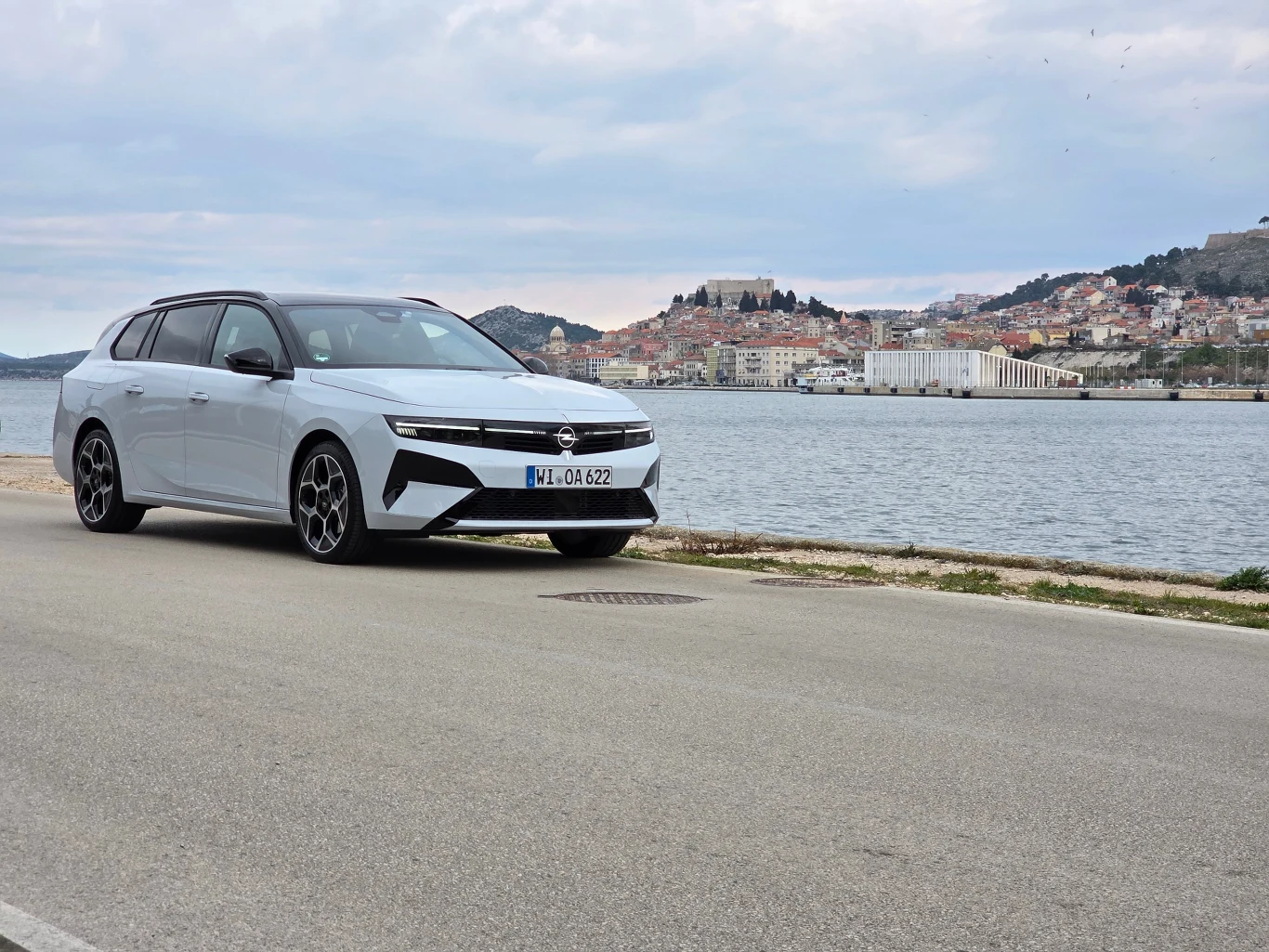biały samochód osobowy stoi zaparkowany przy nabrzeżu, w tle widoczna zabudowa miasta na wzgórzach oraz fragment akwenu wodnego