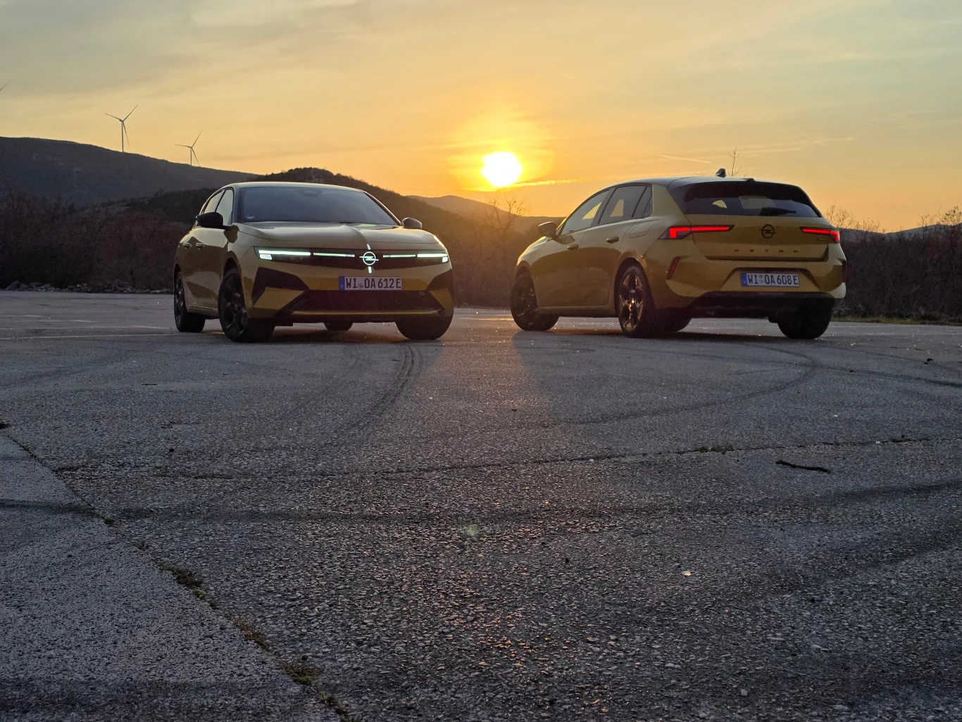 Dwa żółte samochody marki Opel ustawione na pustym, asfaltowym placu, na tle zachodzącego słońca i wzgórz, z widocznymi charakterystycznymi reflektorami i nowoczesną sylwetką pojazdów.