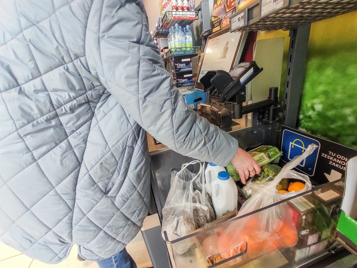 osoba w szarej, pikowanej kurtce układa zakupy na taśmie przy kasie samoobsługowej; w koszyku znajdują się warzywa, mleko oraz inne produkty spożywcze