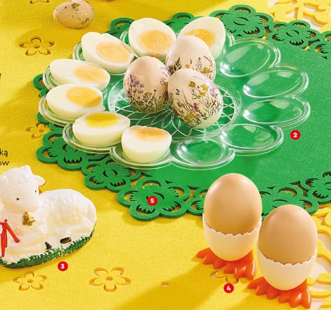 plasikowa patera z zielonym ażurowym zdobieniem z ułożonymi na niej połówkami jajek na twardo oraz ozdobnymi jajkami z kwiatowym motywem, obok baranek z cukru oraz dwa jajka w skorupkach postawione pionowo na pomarańczowych podstawkach w kształcie łapek