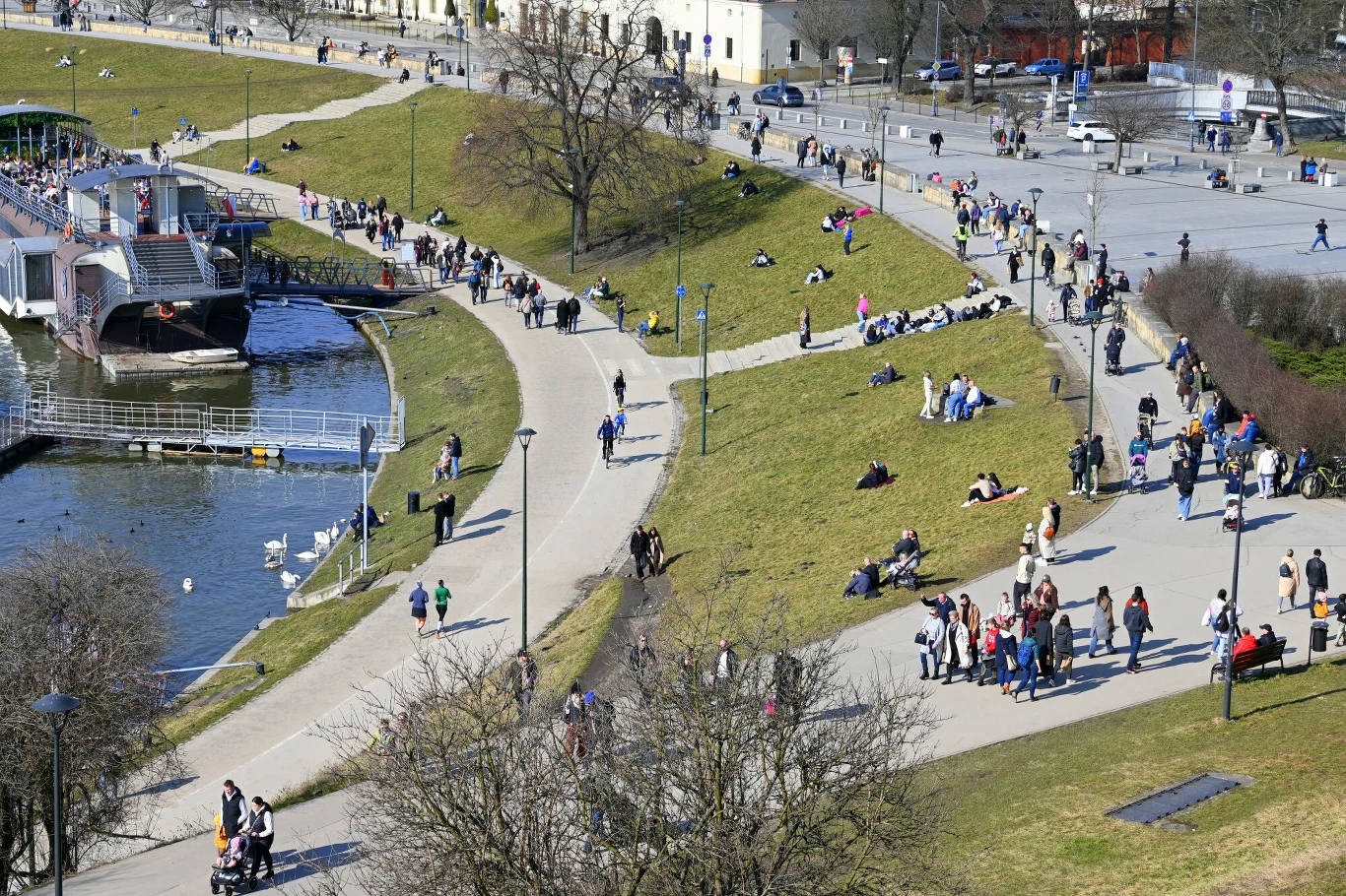 duża liczba spacerujących i odpoczywających osób na zielonym bulwarze nad rzeką, tramwaj wodny zacumowany przy nabrzeżu, grupy ludzi na ścieżkach oraz na trawie korzystających z pogody