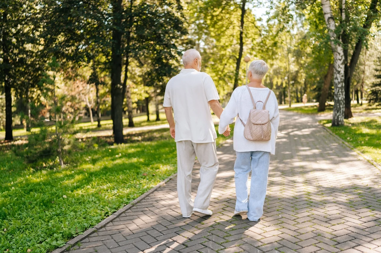 Spacer po posiłku poprawia samopoczucie i jest korzystny dla zdrowia starsza para spacerująca alejką w parku, trzymająca się za ręce, otoczona zielonymi drzewami i słonecznym światłem