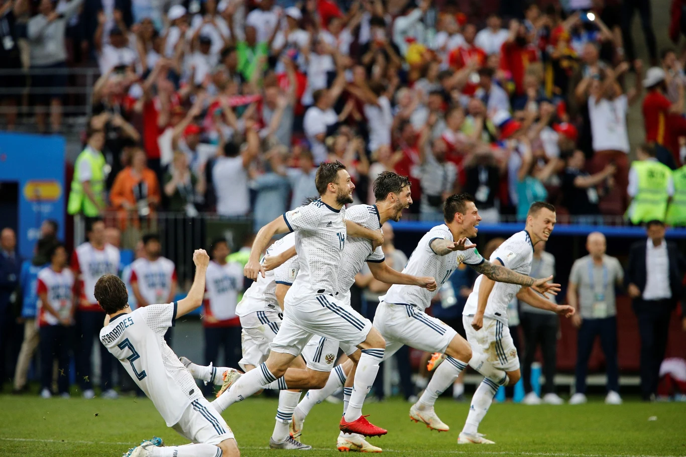 piłkarze w białych strojach biegnący i świętujący na boisku po odniesieniu zwycięstwa, w tle tłum wiwatujących kibiców na trybunach