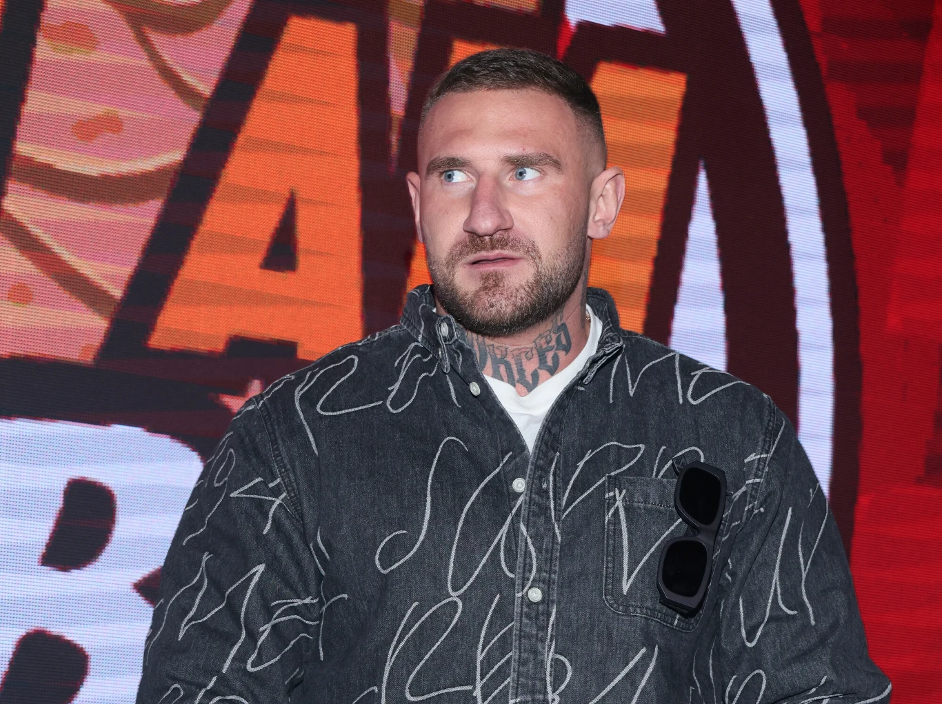 Mężczyzna z krótkimi włosami i brodą, mający tatuaż na szyi, ubrany w ciemną koszulę z jasnym wzorem i ciemne okulary zawieszone na kieszeni, stoi na tle dużego, kolorowego napisu na ścianie.