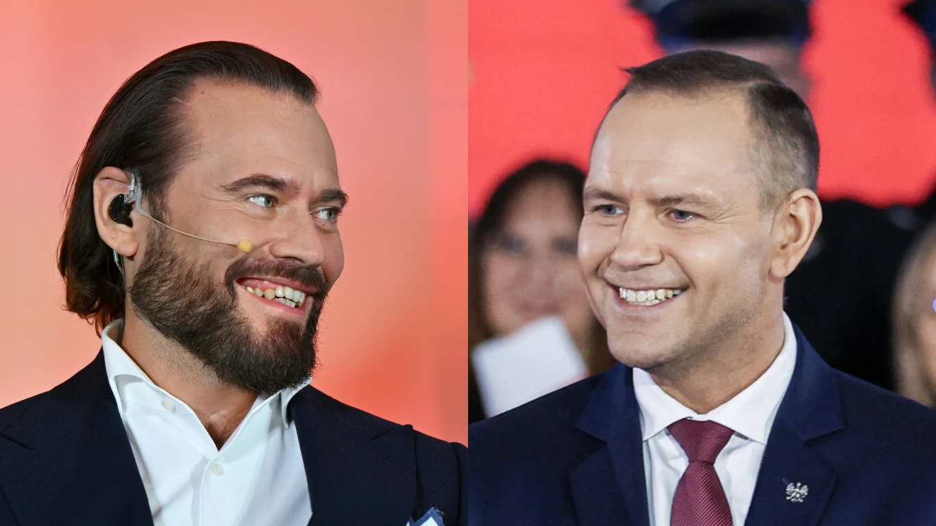 Dwóch mężczyzn ubranych w garnitury uśmiecha się do siebie, jeden ma słuchawkę na uchu i zarost, drugi jest gładko ogolony, w tle rozmyte postacie i ciepłe, czerwone światło.