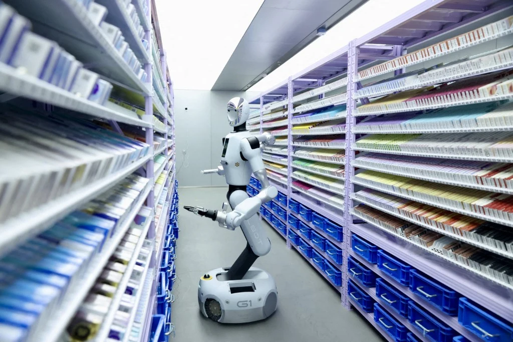 robot humanoidalny o białej obudowie poruszający się pomiędzy regałami z lekami w nowoczesnym magazynie farmaceutycznym, z półkami zorganizowanymi według rodzajów produktów