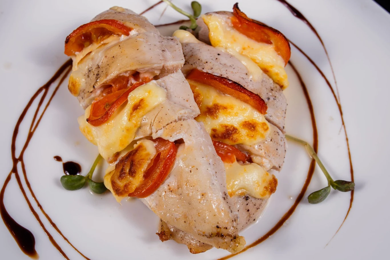 Zapiekany filet z kurczaka idealnie sprawdzi się na obiad Zapiekany filet z kurczaka z plastrami pomidora i sera, podany z sosem balsamicznym i kiełkami na talerzu.