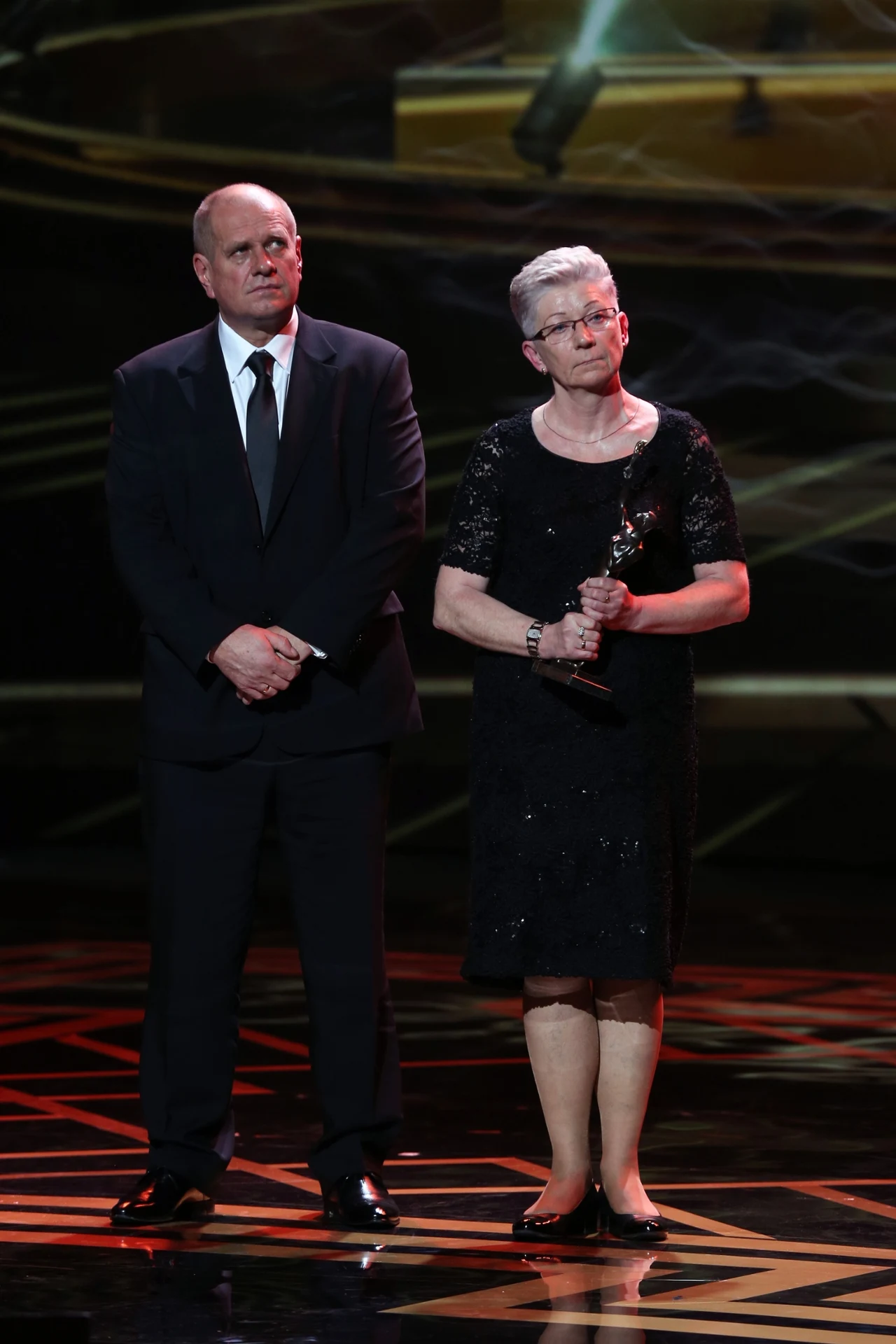 Kobieta w czarnej sukience trzymająca statuetkę stoi obok mężczyzny w ciemnym garniturze na scenie z eleganckim, złotym tłem i reflektorami skierowanymi na nich.