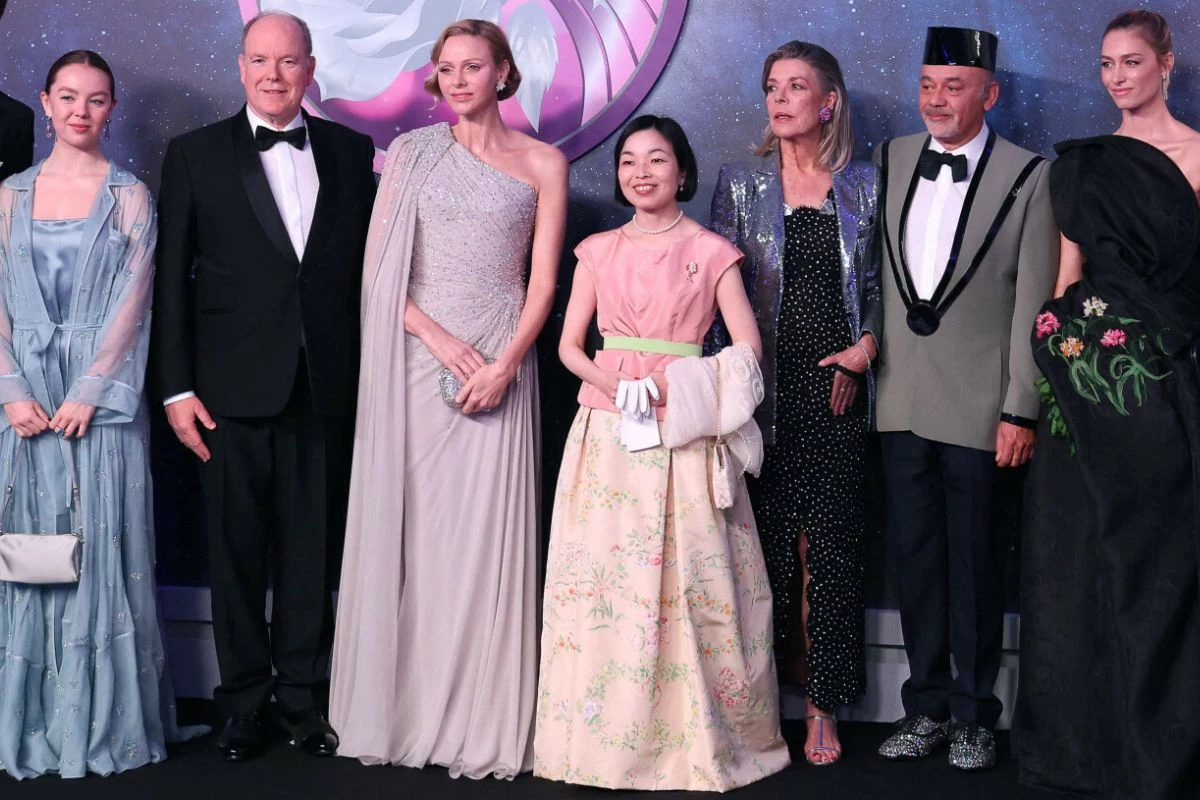 grupa elegancko ubranych osób pozuje na tle dekoracji w stylu galowym, większość kobiet ma na sobie długie suknie, a mężczyźni noszą smokingi lub fraki, centralne miejsce zajmuje kobieta w jasnoszarej sukni stojąca obok mężczyzny w czarnym smokingu