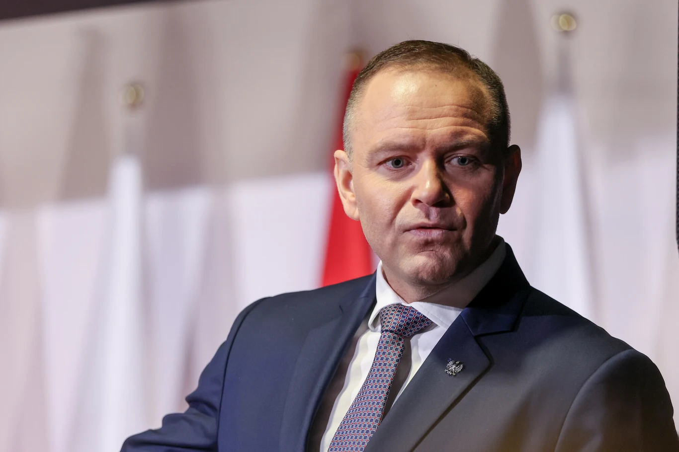 Mężczyzna w garniturze i krawacie stoi na tle biało-czerwonych flag, patrzy lekko w bok z poważnym wyrazem twarzy.