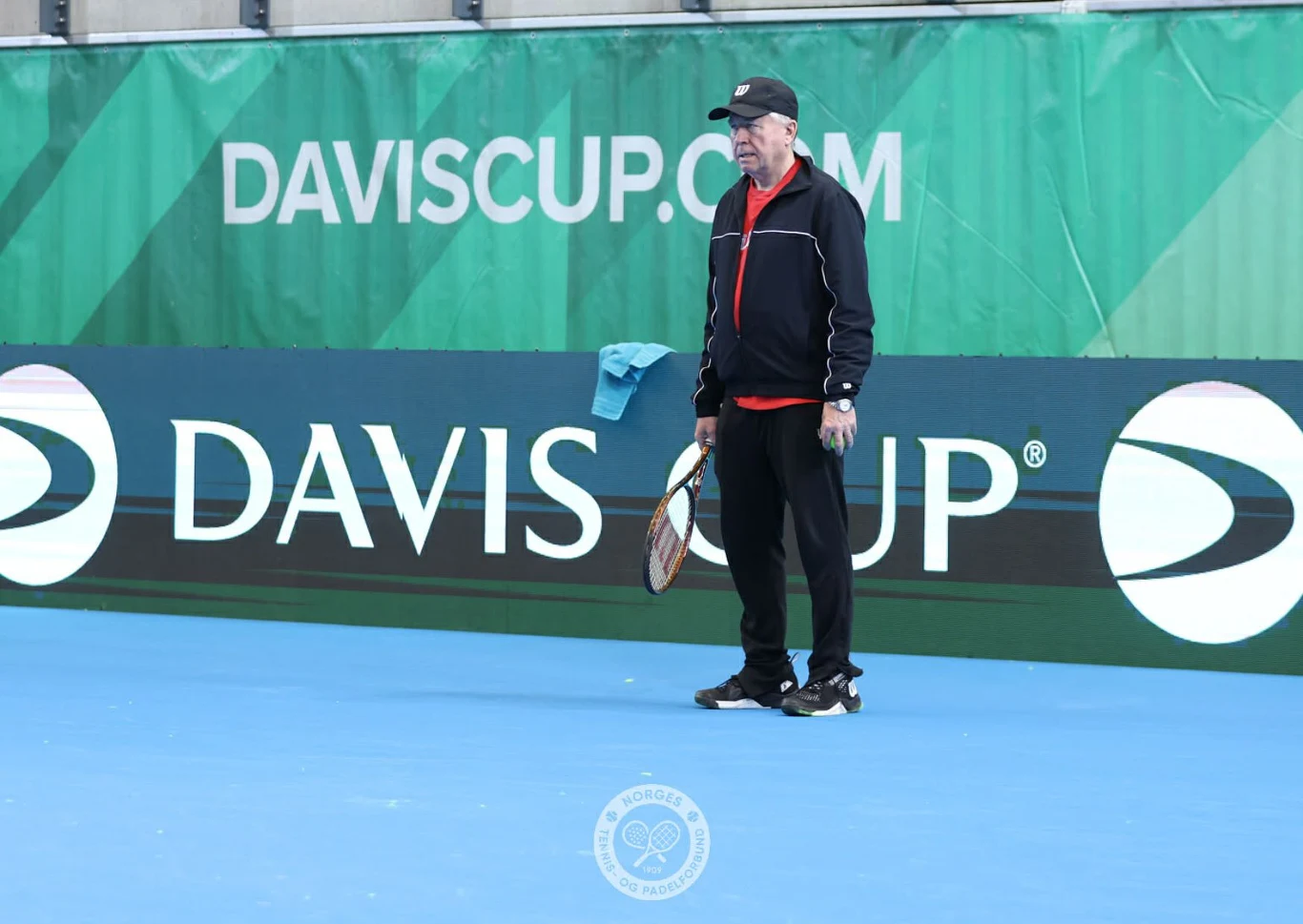 mężczyzna w czarnej sportowej odzieży i czapce stoi na kortcie tenisowym, trzymając rakietę w dłoni, na tle zielonego baneru z napisem 'DAVISCUP.COM'