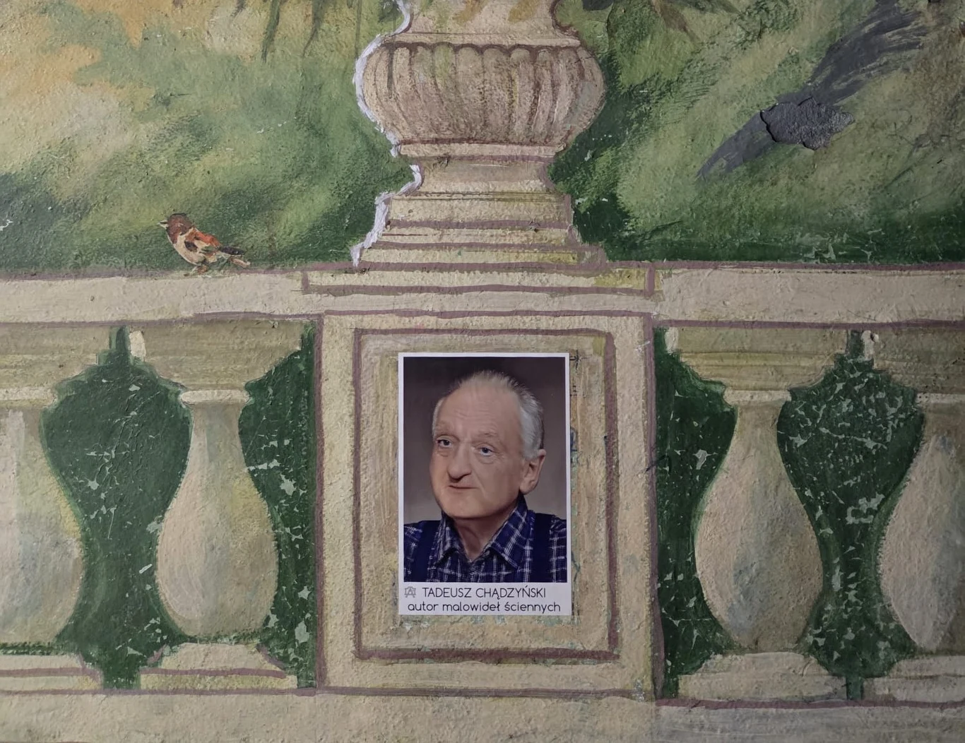 portret starszego mężczyzny z siwymi włosami i łagodnym wyrazem twarzy, umieszczony w ozdobnej, namalowanej ramie imitującej mur z balustradą; po lewej stronie na balustradzie siedzi niewielki ptak, w tle zielone liście i fragment kamiennej wazy