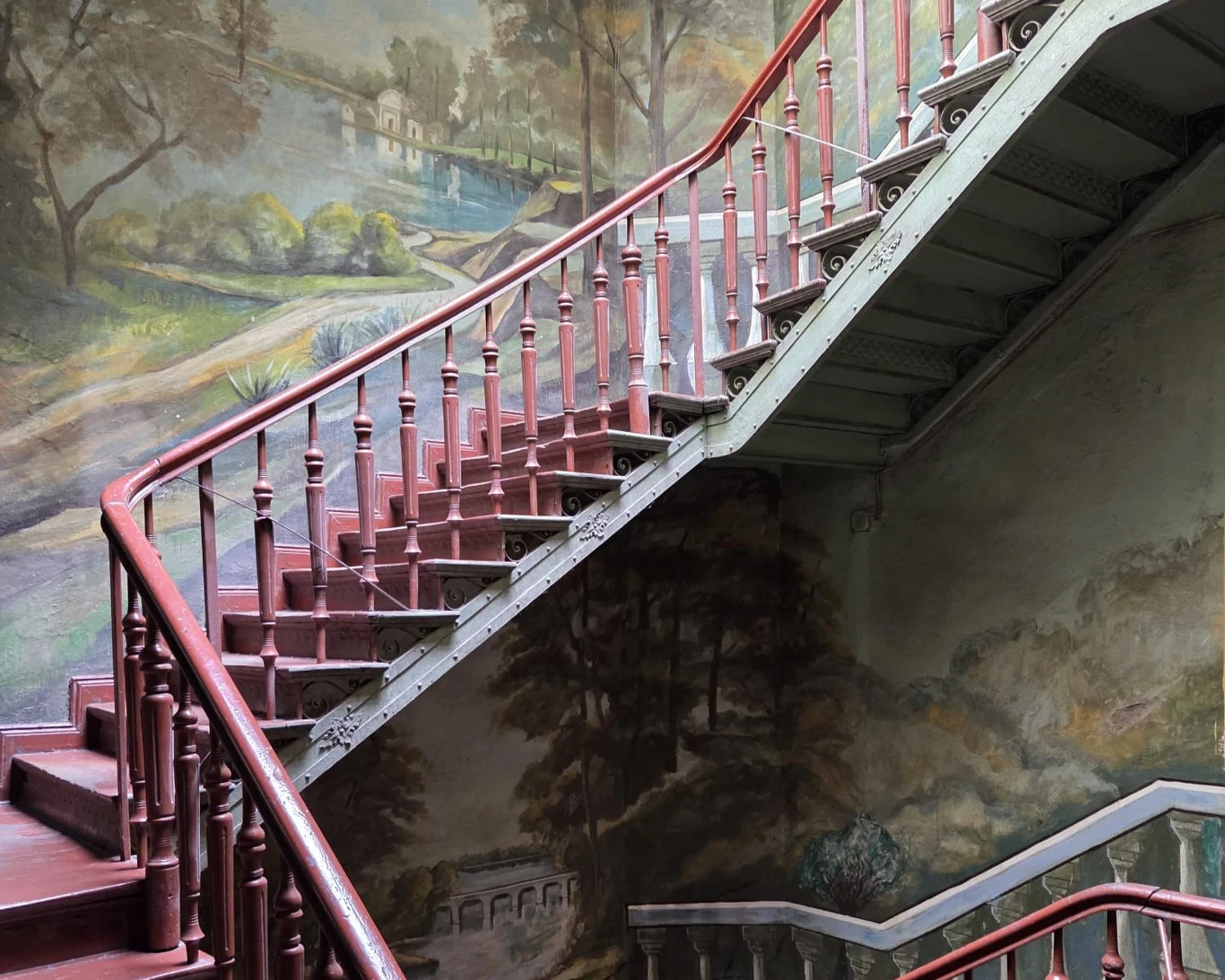 czerwone drewniane schody z balustradą prowadzące w górę, otoczone ścianami ozdobionymi malowidłami przedstawiającymi krajobraz z drzewami i wiejskim domem w tle