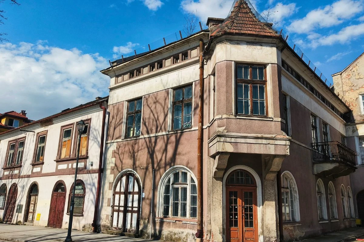 stara kamienica o jasnobrązowej fasadzie z dużymi oknami, balkonem z kutą balustradą oraz spiczastym dachem, otoczona nagimi drzewami, których cienie padają na ścianę budynku