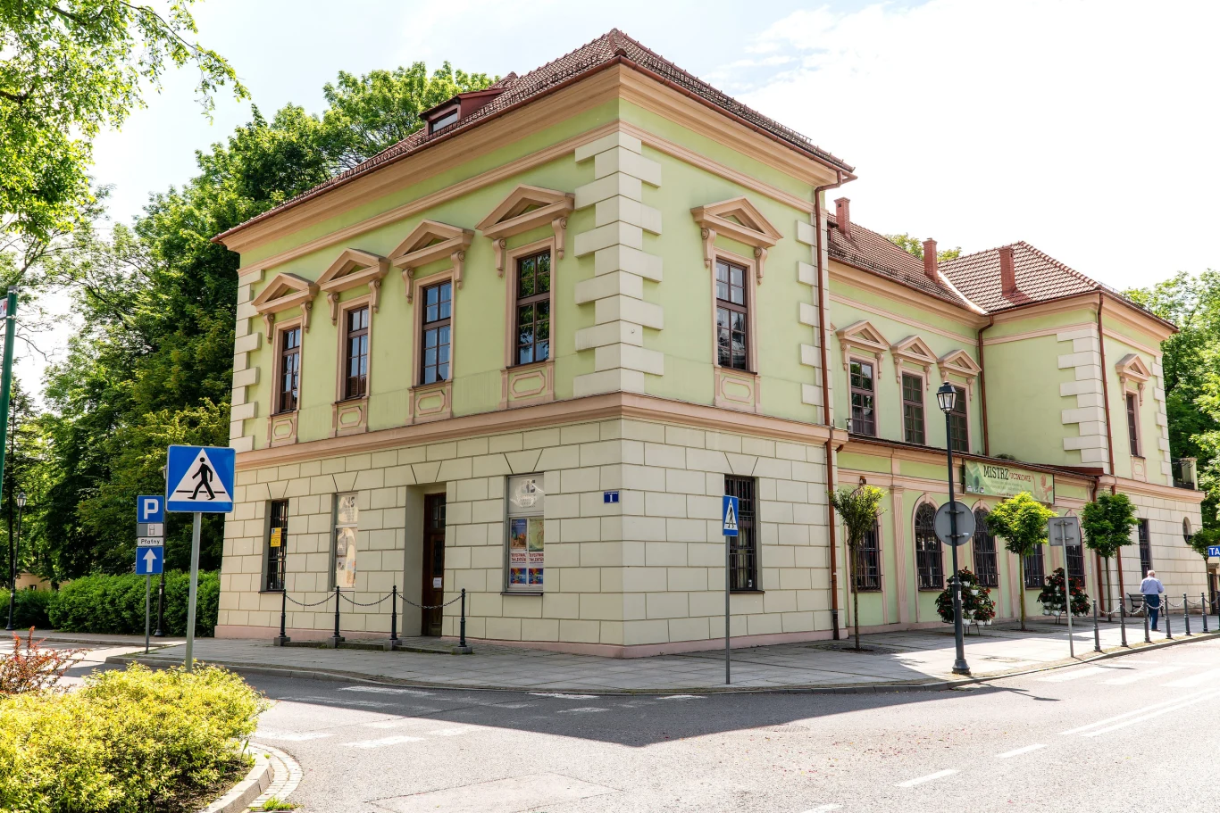 Dwupiętrowy, jasny budynek w klasycznym stylu z wysokimi oknami i ozdobnymi gzymsami, stojący na rogu ulic otoczonych zielenią, z widocznymi znakami drogowymi, w tym przejściem dla pieszych i tablicami parkingowymi.