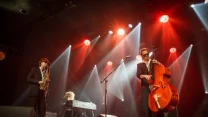 Trzech muzyków grających na scenie: saksofonista, kontrabasista oraz pianista, oświetleni intensywnymi reflektorami, wszyscy ubrani elegancko na czarno, atmosfera koncertowa z wyraźnym skupieniem na muzyce.
