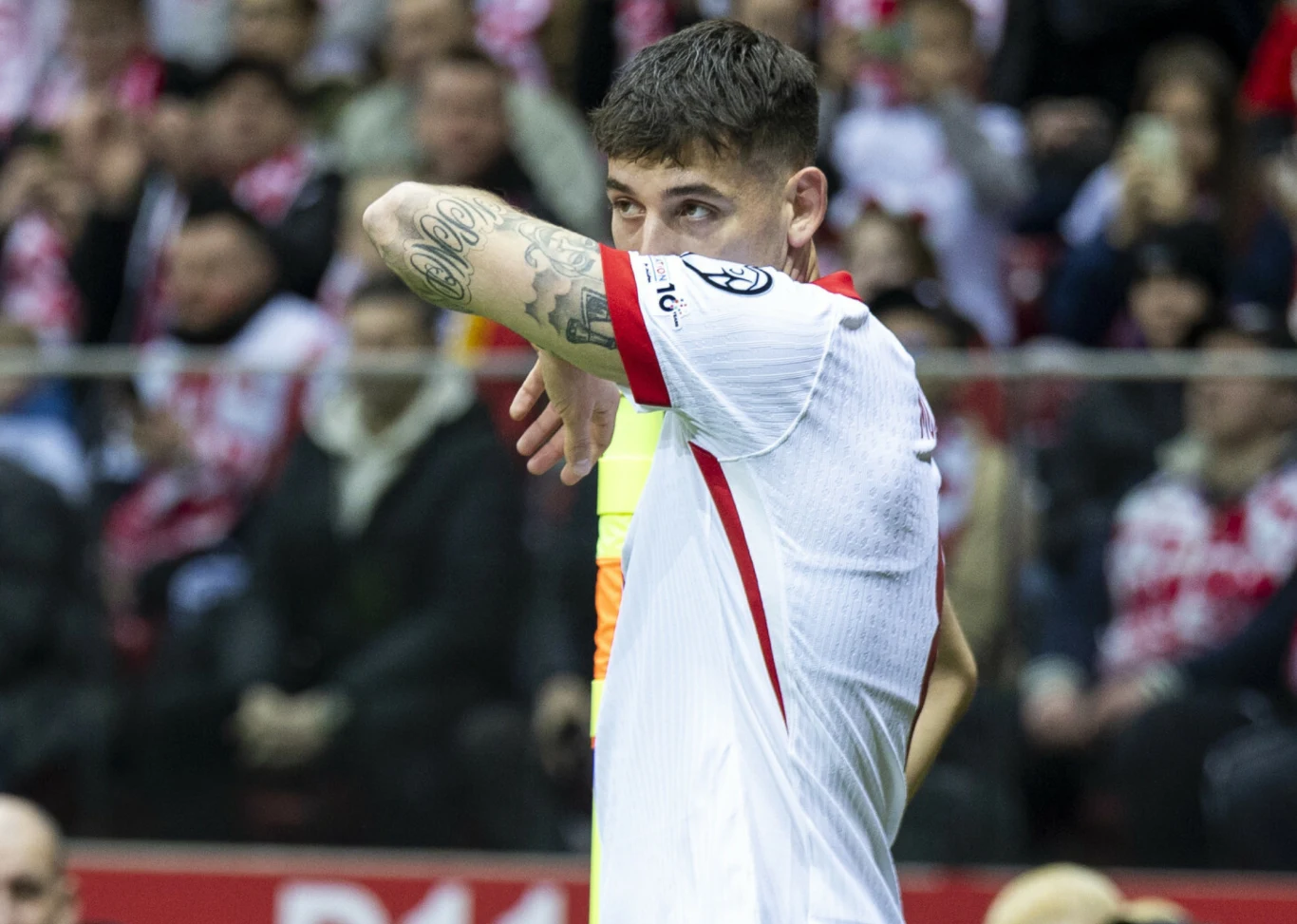 piłkarz w białej koszulce z czerwonymi akcentami ociera twarz przedramieniem, prezentując tatuaż na lewej ręce; w tle rozmyci kibice na trybunach