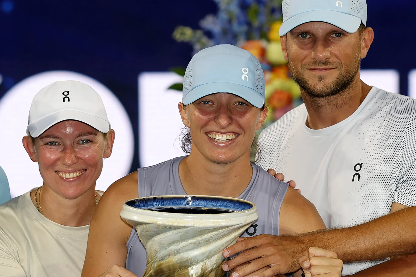 uśmiechnięta kobieta trzyma duże trofeum, obok niej stoją dwie osoby, wszyscy mają na sobie jasne koszulki i czapki z jednym logo, w tle widać rozmyte kolorowe kwiaty
