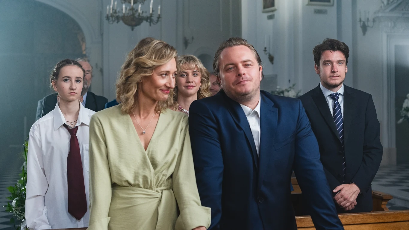 grupa pięciu osób ubranych elegancko, stojących w kościele; na pierwszym planie kobieta w jasnej sukience i mężczyzna w granatowym garniturze, wyglądają na zadowolonych, w tle trzy młode osoby, z których jedna wyróżnia się luźnym strojem i dużym krawatem
