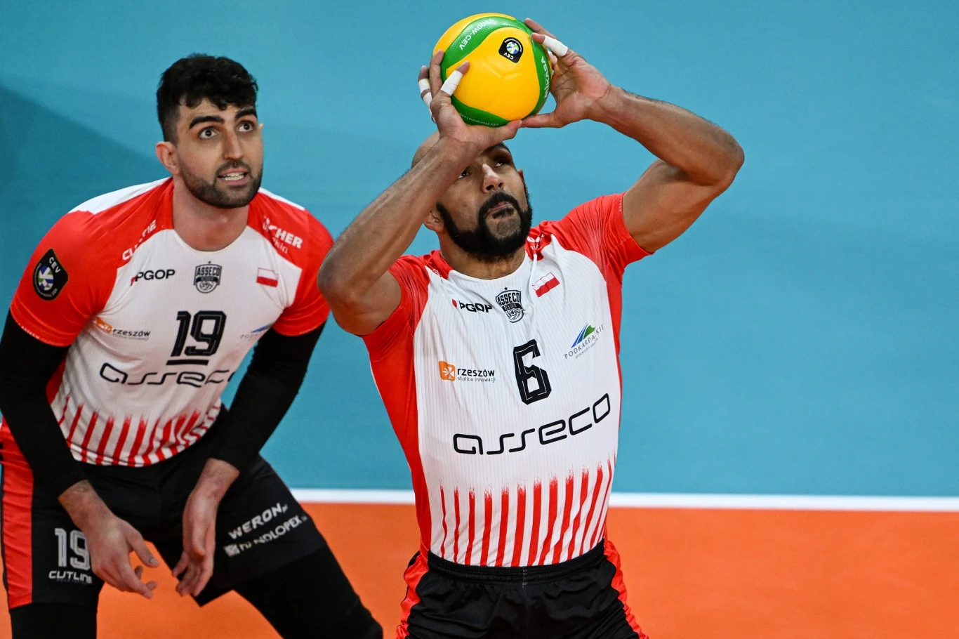 Dwóch siatkarzy w biało-czerwonych koszulkach podczas akcji – jeden z nich koncentruje się na precyzyjnym rozegraniu piłki na wysokości głowy, drugi obserwuje i przygotowuje się do kolejnego ruchu na boisku.