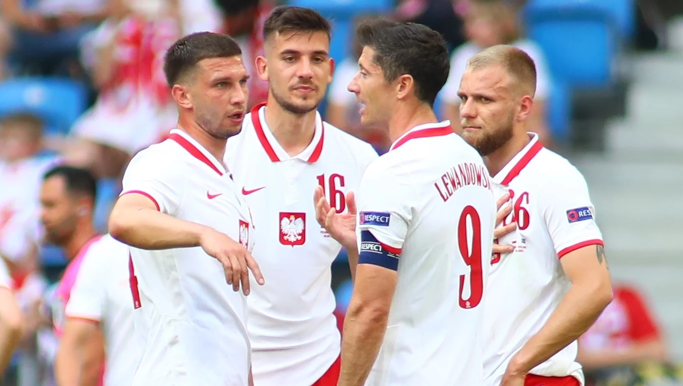 Czterech piłkarzy w biało-czerwonych strojach reprezentacji Polski stoi blisko siebie na boisku, prowadząc żywiołową rozmowę podczas meczu, w tle widoczne niebieskie krzesełka stadionu.