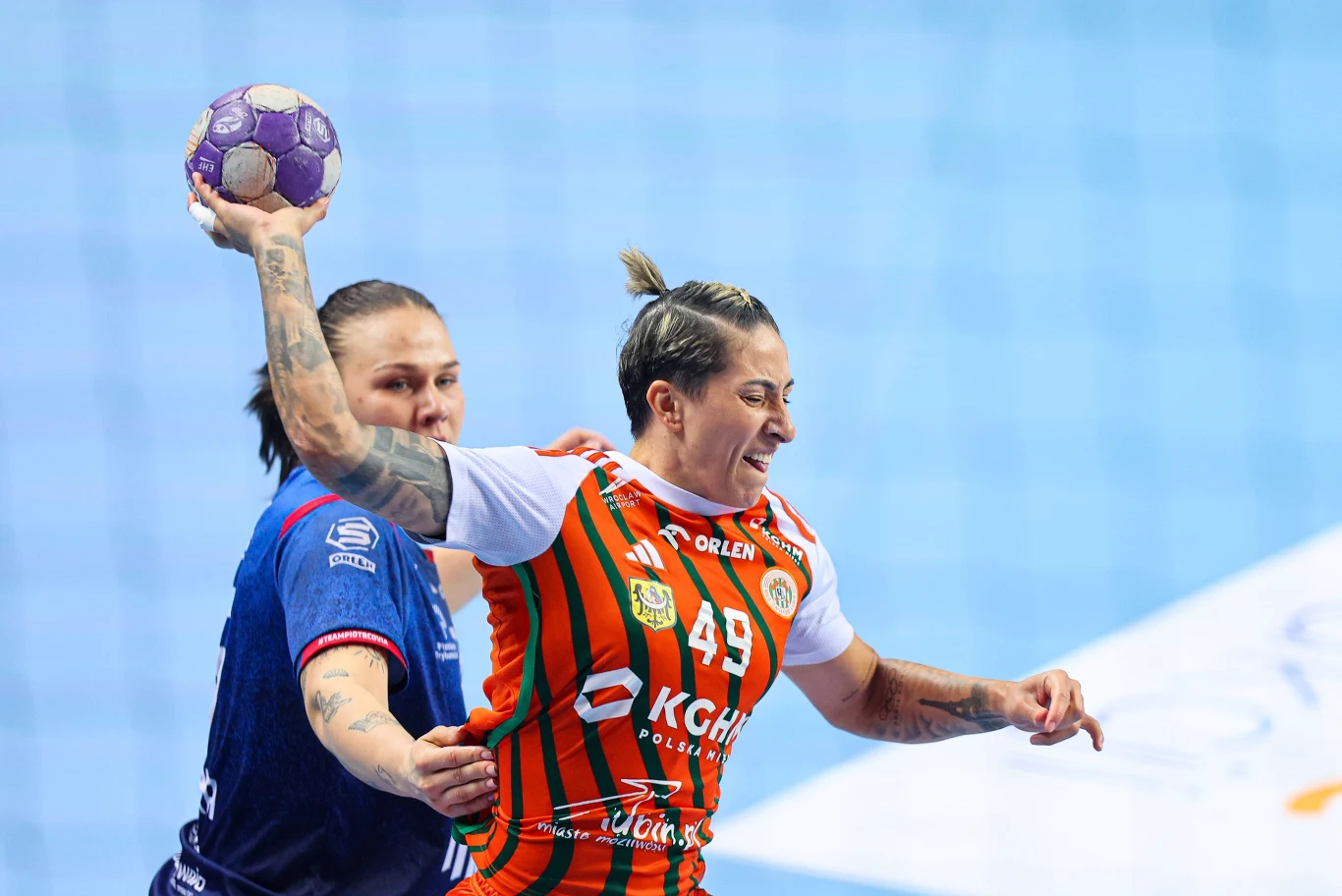 zawodniczka w pomarańczowo-zielonej koszulce z uniesioną ręką trzyma piłkę do piłki ręcznej, w tle widoczna przeciwniczka próbująca ją zatrzymać podczas dynamicznej akcji na boisku
