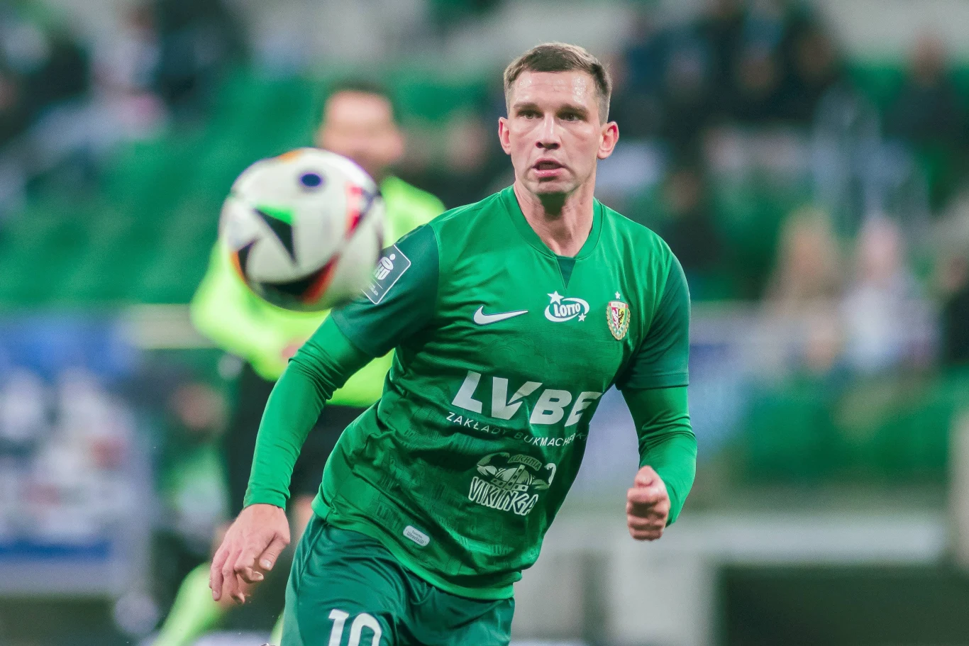 piłkarz w zielonym stroju Śląska Wrocław biegnie na boisku, skupiony na zbliżającej się piłce podczas meczu