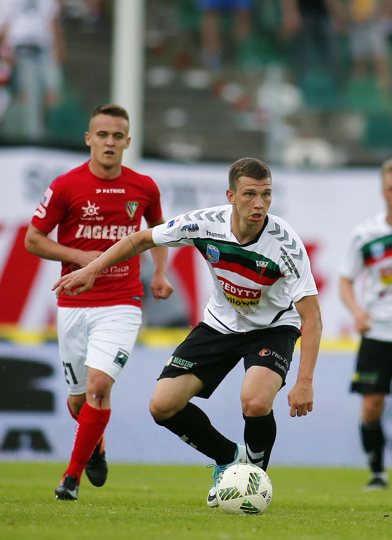 piłkarz w biało-czarnym stroju prowadzi piłkę podczas meczu piłkarskiego, a za nim znajduje się zawodnik w czerwonej koszulce