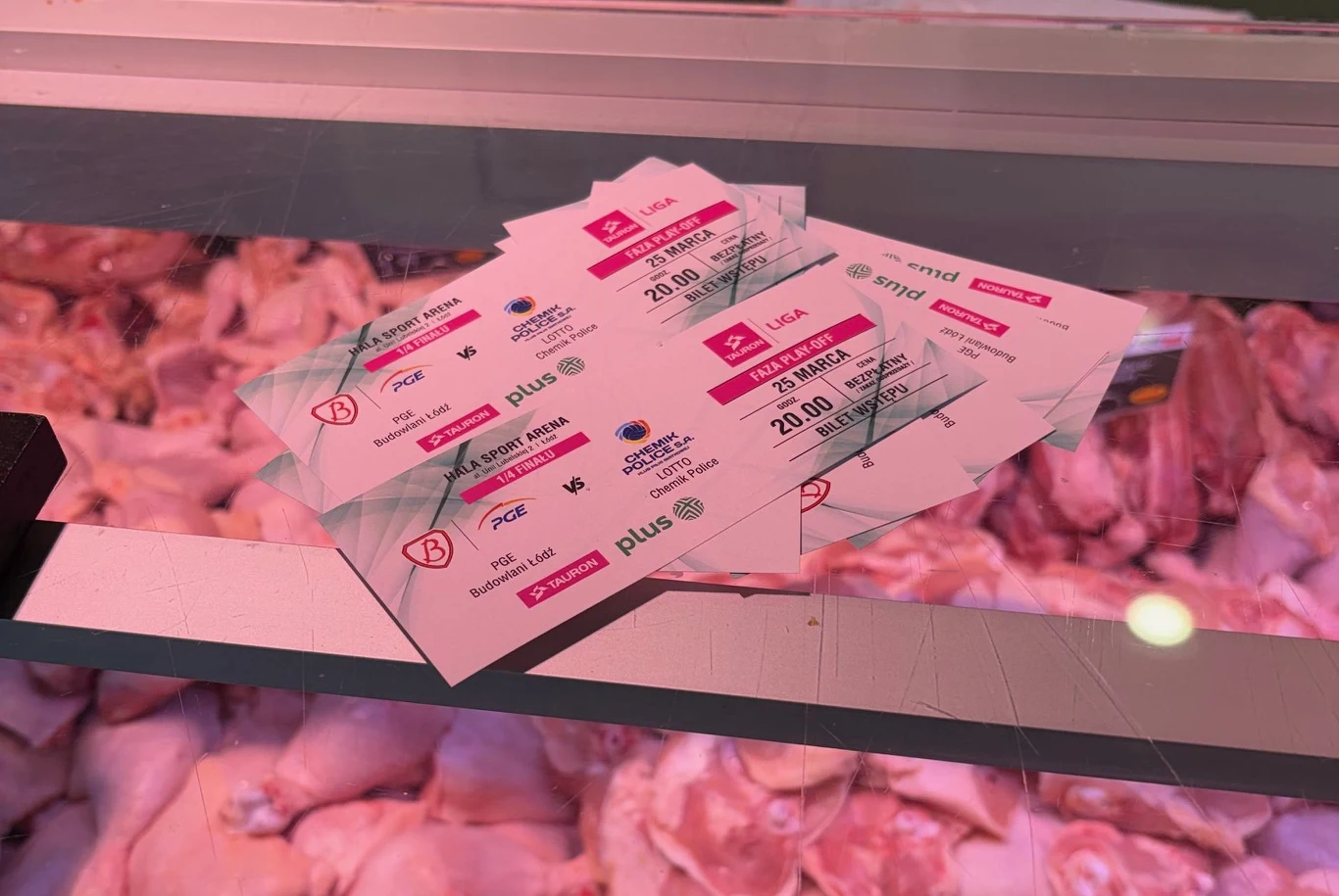 kilka papierowych biletów lub wejściówek z nadrukowanymi informacjami leży na ladzie chłodniczej z surowym mięsem, na pierwszym planie dominują jasne odcienie mięsa i wyraźne, drukowane napisy na kartkach