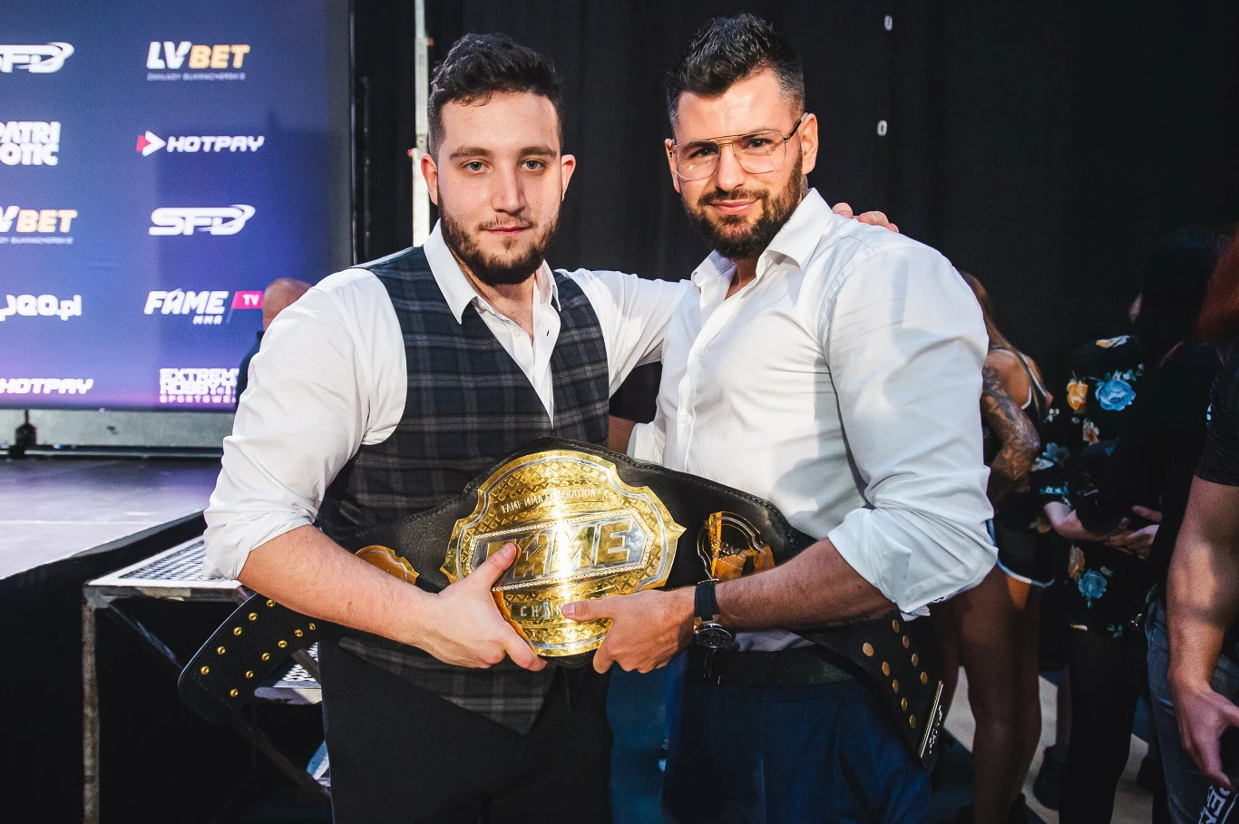 dwóch mężczyzn stoi razem obejmując się, jeden z nich trzyma dużą, złotą mistrzowską pasę, obaj ubrani elegancko, w tle ściana z logo sponsorów