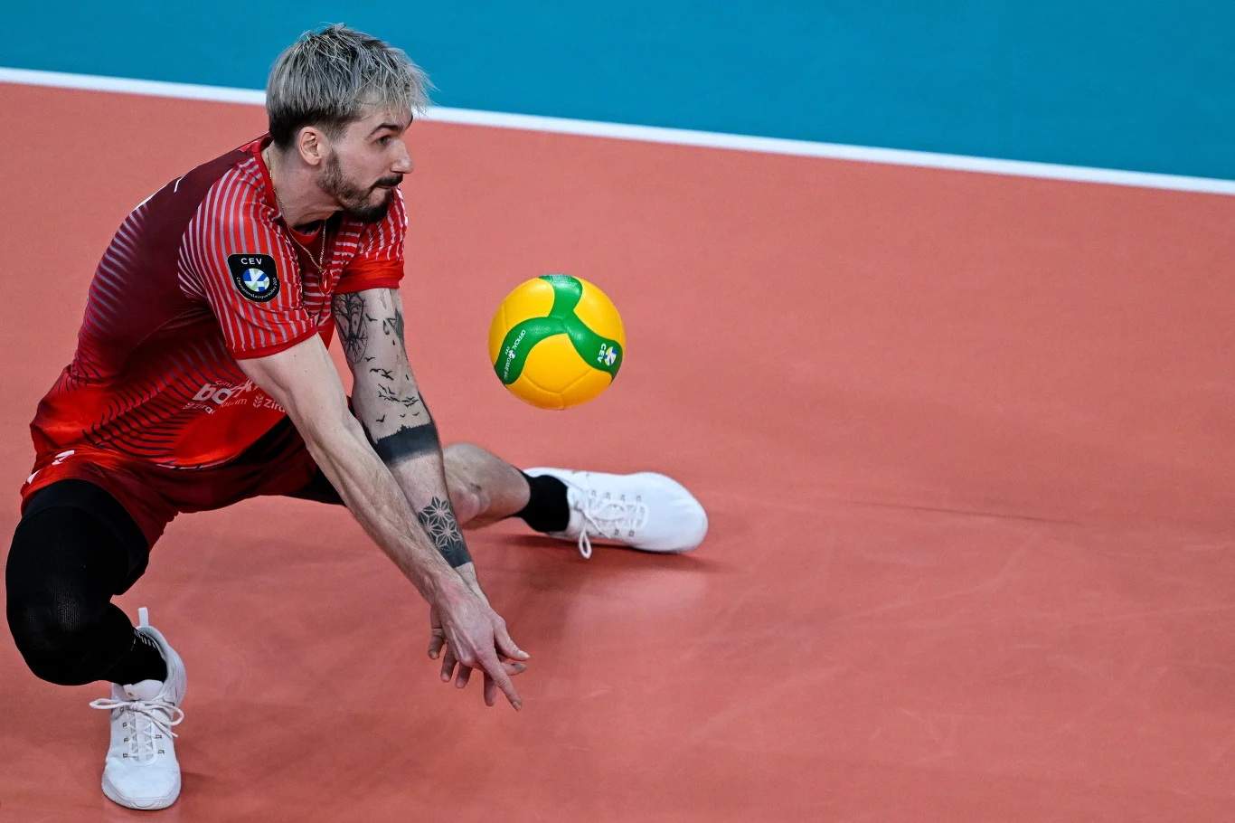 siatkarz w czerwonym stroju wykonujący odbiór piłki sposobem dolnym na hali sportowej, skoncentrowany na precyzyjnym przyjęciu, piłka tuż przed dłońmi zawodnika
