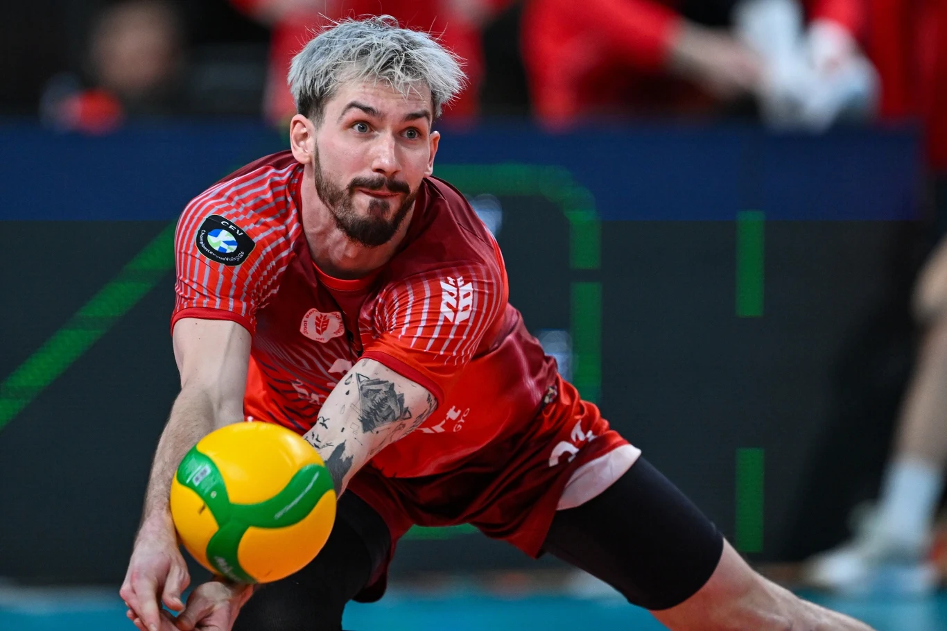 siatkarz w czerwonym stroju dynamicznie odbija piłkę tuż nad ziemią, skupienie widoczne na twarzy, na ramieniu duży tatuaż