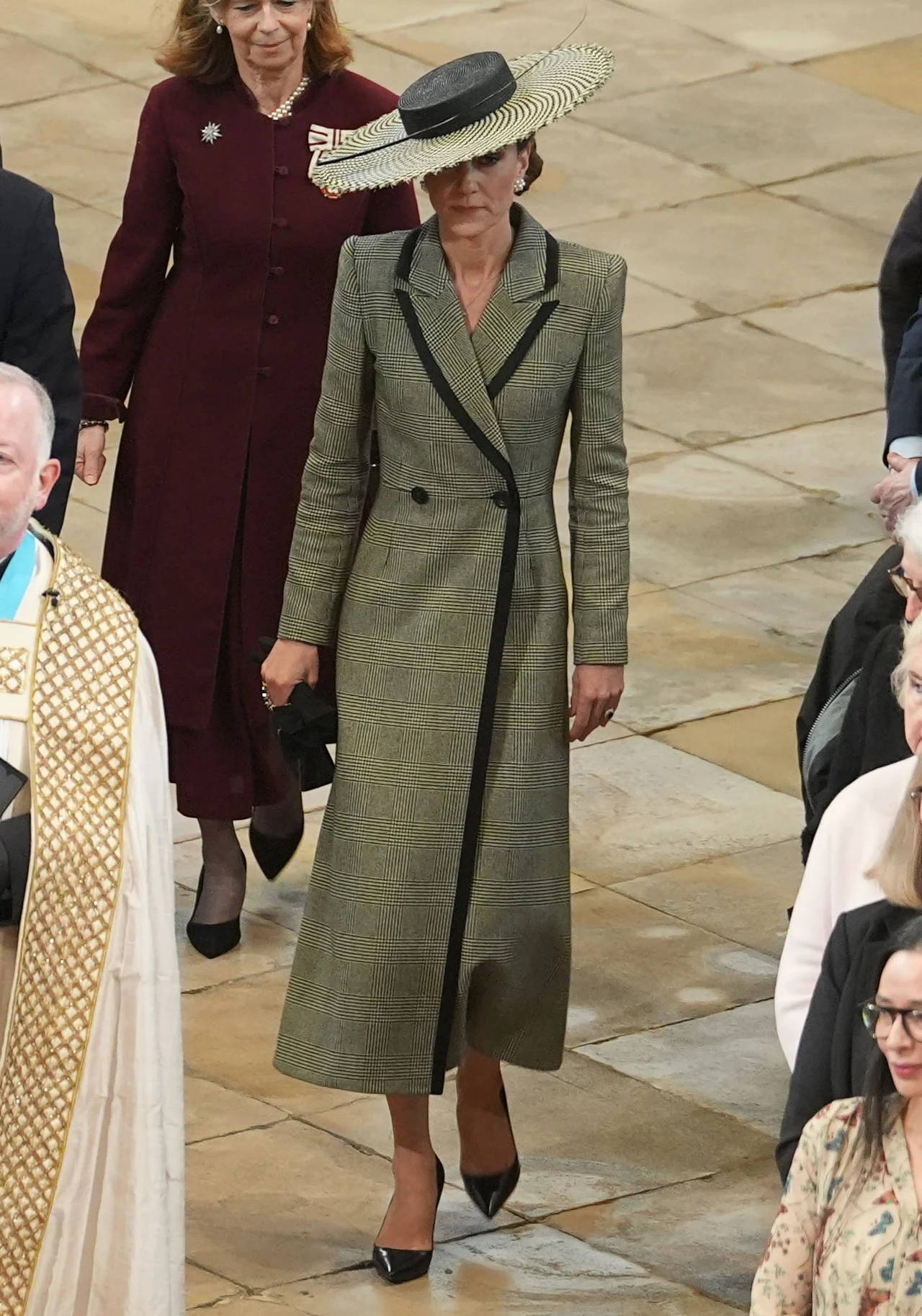 kobieta ubrana w długi, klasyczny zielony płaszcz z czarnymi akcentami i czarnymi guzikami, na głowie ma duży kapelusz z szerokim, kontrastowym rondem; stoi na kamiennej posadzce, wokół niej znajdują się inne elegancko ubrane osoby