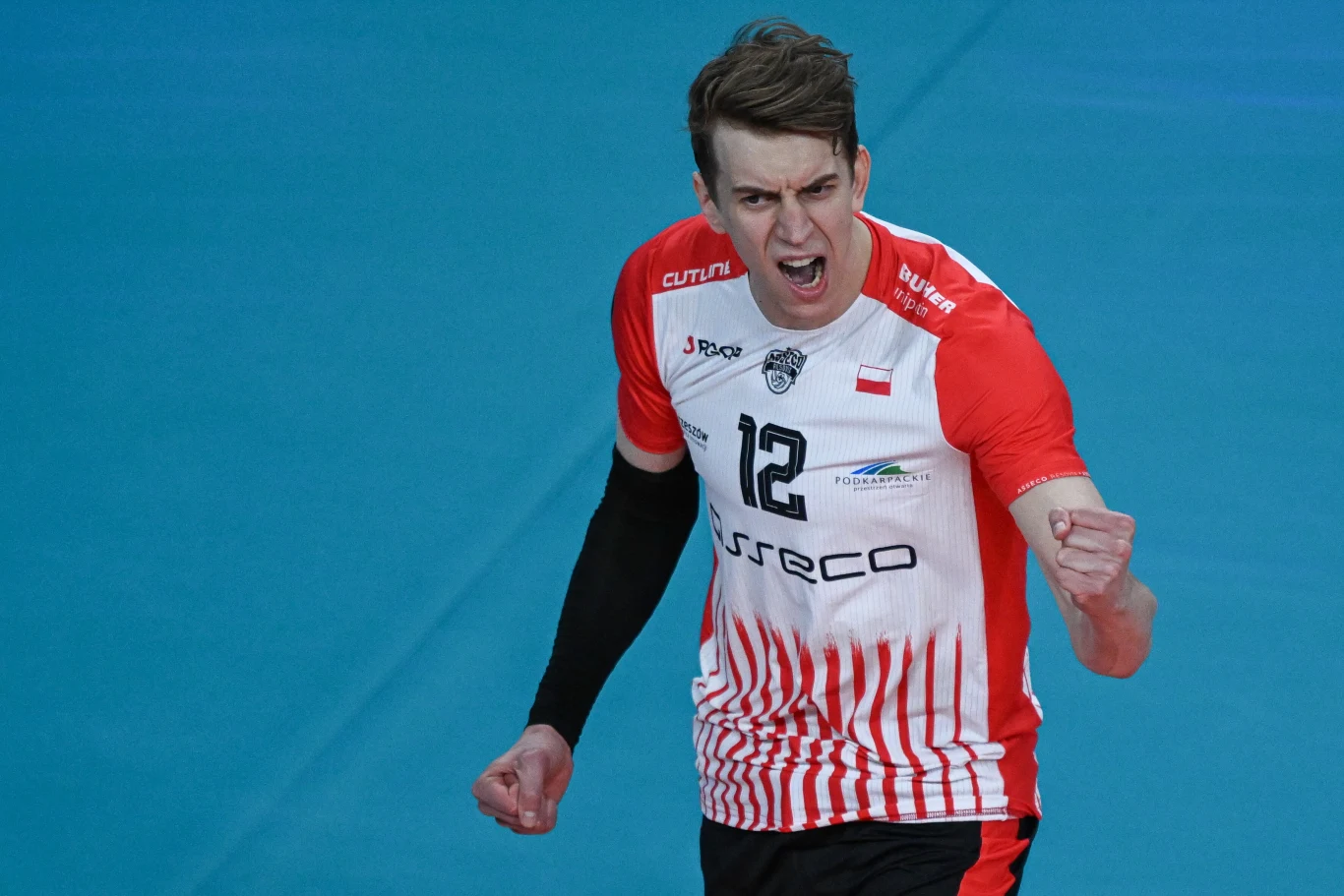 siatkarz w czerwono-białym stroju z numerem 12 na koszulce świętuje zdobycie punktu, zaciskając pięści i wyrażając silne emocje na tle niebieskiego boiska