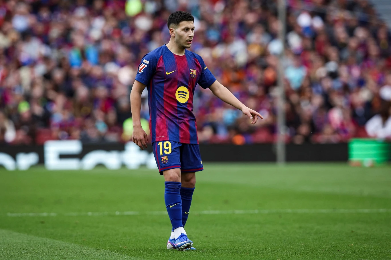 piłkarz w granatowo-bordowym stroju FC Barcelona z numerem 19 na plecach stoi na murawie stadionu, wokół widoczna rozmyta publiczność